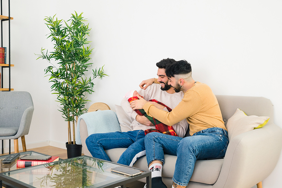 smiling-young-gay-couple-sitting-sofa-loving-their-baby-900 Smiling gay couple loving baby